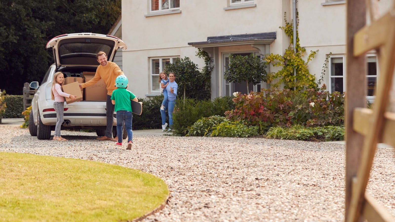 A family of five unloads boxes on the gravel driveway of their new home as the son with a skateboard runs to help.