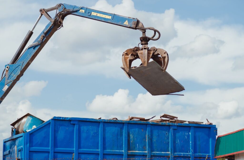 A crane is dropping debris into a blue dumpster. 