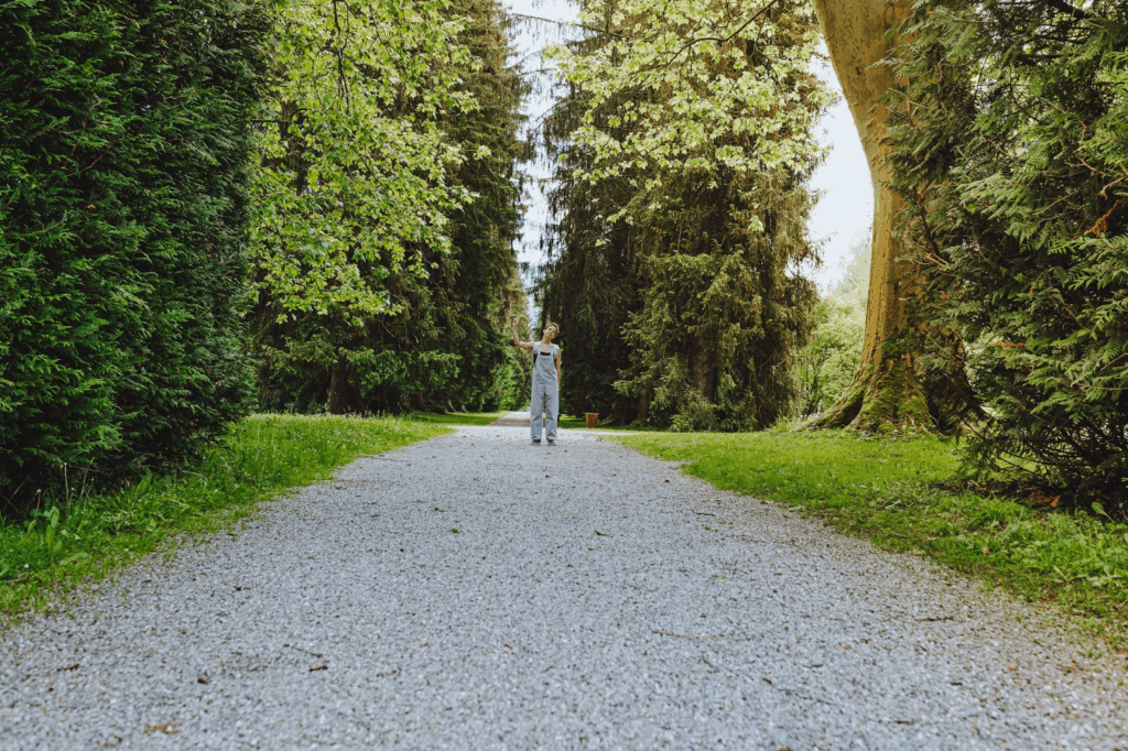 A girl stands in the middle of a gravel driveway surrounded by tall trees.