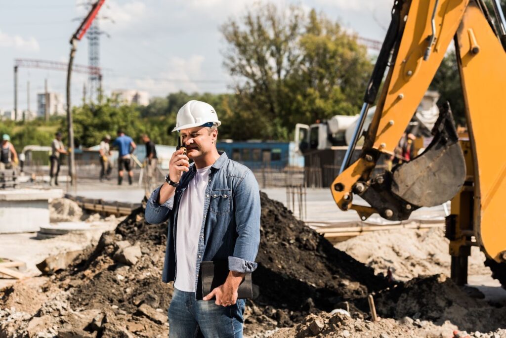 A demolition contractor stands on a jobsite and talks on a walkie talkie.